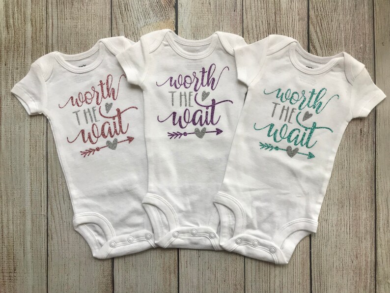 Puede incluir: Tres bodies blancos para beb&eacute; con el texto "worth the wait" en letras de purpurina. Cada body tiene un color de purpurina diferente: rosa, morado y turquesa.