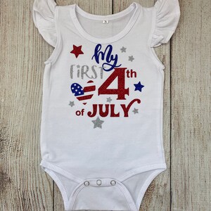 May include: White baby onesie with red, white, and blue glitter text that reads "My First 4th of July". The text is surrounded by silver glitter stars.