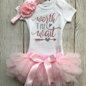 Puede incluir: Un tut&uacute; rosa con un lazo de sat&eacute;n rosa, un body blanco con el texto "worth the wait" en purpurina plateada y rosa, y una diadema rosa con una flor rosa.