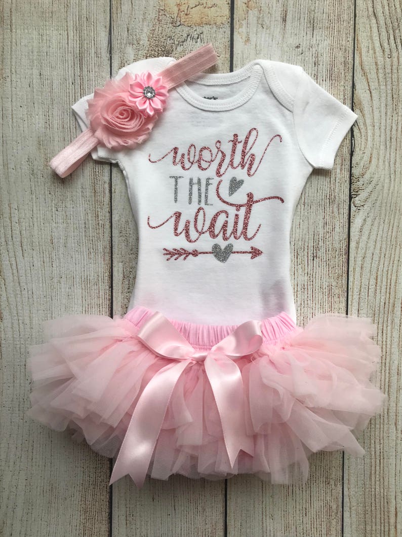 Puede incluir: Un tut&uacute; rosa con un lazo de sat&eacute;n rosa, un body blanco con el texto "worth the wait" en purpurina plateada y rosa, y una diadema rosa con una flor rosa.