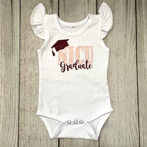 May include: White baby onesie with a pink and maroon graphic that says "NICU Graduate" with a graduation cap above the text.