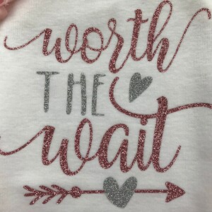 Puede incluir: Un body blanco con el texto "worth the wait" en purpurina rosa y plateada. El texto est&aacute; rodeado por un coraz&oacute;n de purpurina plateada y una flecha con un coraz&oacute;n de purpurina plateada en el medio.