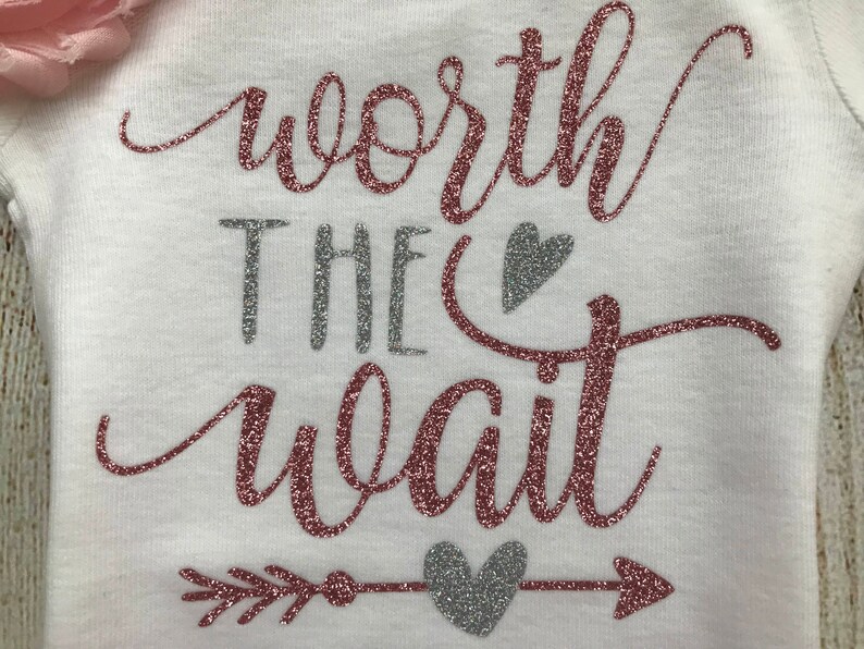 Puede incluir: Un body blanco con el texto "worth the wait" en purpurina rosa y plateada. El texto est&aacute; rodeado por un coraz&oacute;n de purpurina plateada y una flecha con un coraz&oacute;n de purpurina plateada en el medio.