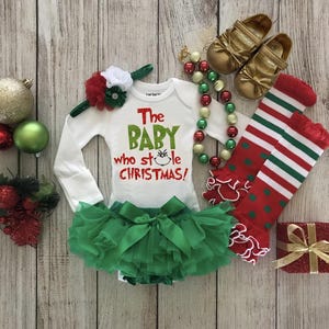 May include: A white onesie with the text "The Baby Who Stole Christmas!" in green and red letters. The onesie is paired with a green tulle tutu, a red and white headband, a green and red beaded necklace, red and green striped knee-high socks, and gold shoes.