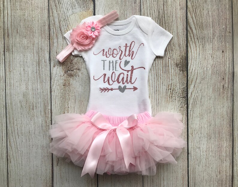 Puede incluir: Un tut&uacute; rosa con un body blanco que dice "worth the wait" en purpurina dorada rosa con una flecha y un coraz&oacute;n en purpurina plateada. El body tiene una diadema de flores rosas con un strass.