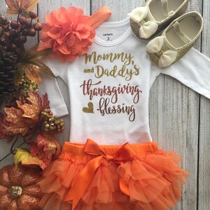 Peut inclure: Tenue de Thanksgiving orange et or pour un b&eacute;b&eacute;. La tenue comprend un body blanc avec le texte "Mommy and Daddy's Thanksgiving Blessing", une jupe en tulle orange avec un n&oelig;ud assorti et un bandeau avec une fleur orange. Le b&eacute;b&eacute; porte &eacute;galement des chaussures &agrave; paillettes dor&eacute;es.