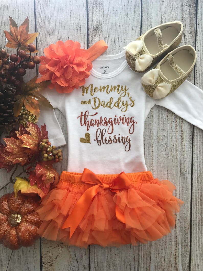 Peut inclure: Tenue de Thanksgiving orange et or pour un b&eacute;b&eacute;. La tenue comprend un body blanc avec le texte "Mommy and Daddy's Thanksgiving Blessing", une jupe en tulle orange avec un n&oelig;ud assorti et un bandeau avec une fleur orange. Le b&eacute;b&eacute; porte &eacute;galement des chaussures &agrave; paillettes dor&eacute;es.