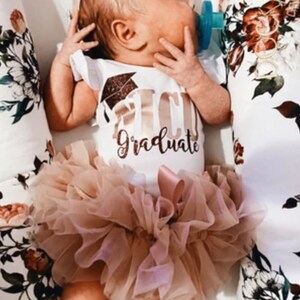 May include: A baby wearing a white onesie with gold glitter text that says "I'm a Graduate" and a gold glitter graduation cap. The baby is also wearing a blush pink tulle tutu.