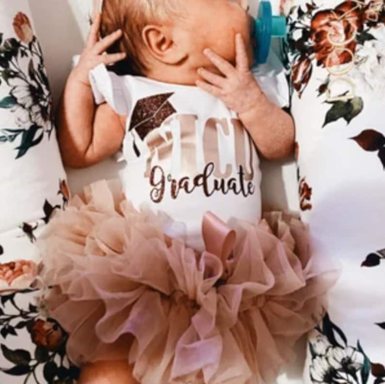 May include: A baby wearing a white onesie with gold glitter text that says "I'm a Graduate" and a gold glitter graduation cap. The baby is also wearing a blush pink tulle tutu.