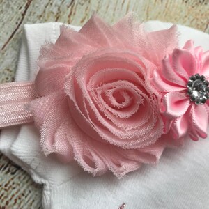 Puede incluir: Una diadema de flores rosa con una gran rosa de tela rosa y una flor de sat&eacute;n rosa m&aacute;s peque&ntilde;a con un centro de pedrer&iacute;a.