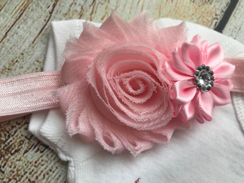 Puede incluir: Una diadema de flores rosa con una gran rosa de tela rosa y una flor de sat&eacute;n rosa m&aacute;s peque&ntilde;a con un centro de pedrer&iacute;a.