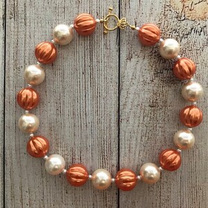 Peut inclure: Un collier compos&eacute; de grosses perles rondes de couleur cr&egrave;me et cuivre. Les perles ont une surface textur&eacute;e, l&eacute;g&egrave;rement bossel&eacute;e. Le collier est ferm&eacute; par un fermoir dor&eacute;.