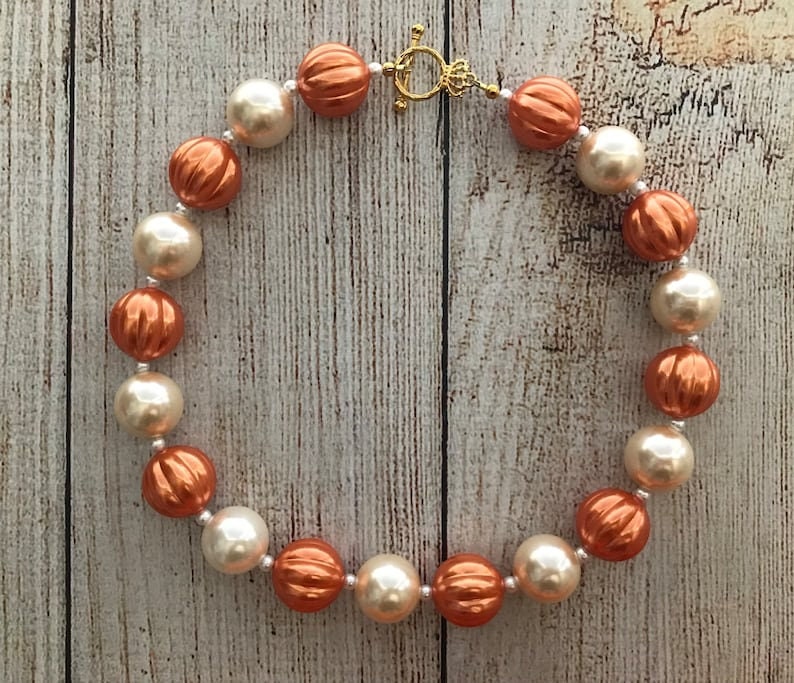 Peut inclure: Un collier compos&eacute; de grosses perles rondes de couleur cr&egrave;me et cuivre. Les perles ont une surface textur&eacute;e, l&eacute;g&egrave;rement bossel&eacute;e. Le collier est ferm&eacute; par un fermoir dor&eacute;.