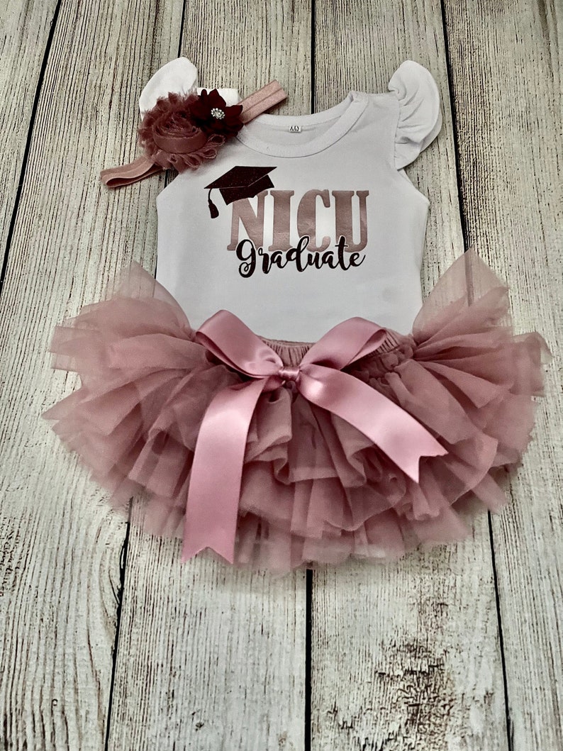 May include: A white onesie with a pink glitter graduation cap and the text "NICU Graduate". The onesie is paired with a pink tulle skirt and a pink headband with a flower.