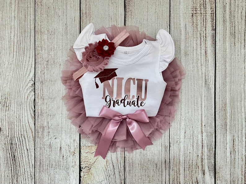 May include: A white onesie with the text "NICU Graduate" in rose gold glitter with a graduation cap graphic. The onesie is paired with a pink tulle tutu and a pink headband with a burgundy flower.