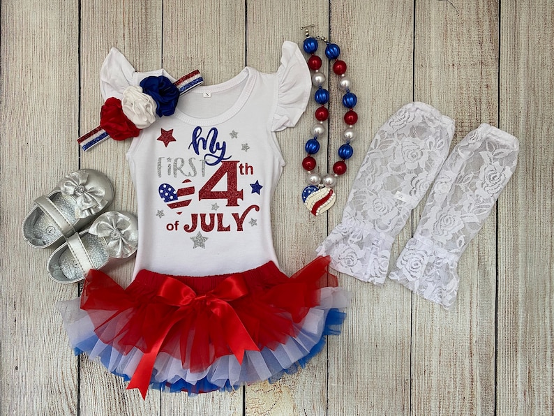 May include: A white bodysuit with red, white, and blue glitter accents. The bodysuit says "My First 4th of July". The outfit includes a red, white, and blue headband, a red, white, and blue tutu, a red, white, and blue beaded necklace, and white lace leg warmers.