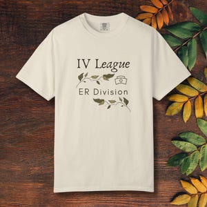 Peut inclure: T-shirt crème avec l'inscription noire "IV League ER Division", accompagnée d'une icône de chapeau d'infirmière et de motifs de branches feuillues. Le t-shirt est en matière douce et a un col rond classique.