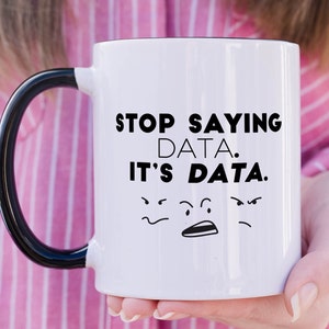 May include: A white ceramic mug with a black handle and interior. The front features the black text "STOP SAYING DATA. IT'S DATA." above three small, hand-drawn black facial expressions showing frustration, confusion, and annoyance with simple lines.