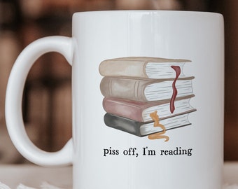 May include: A white ceramic mug featuring a stack of four illustrated books with a red bookmark and a yellow bookmark. Below the books, the text reads, "piss off, I'm reading" in a black serif font. The mug has a curved handle on the left side.