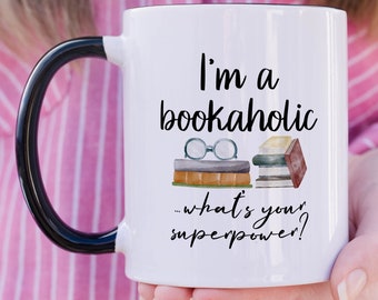 May include: A white ceramic mug with a black handle and rim. The front features the black text 'I'm a bookaholic... what's your superpower?' above a watercolor illustration of a stack of books topped with a pair of round reading glasses.