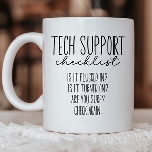May include: A white ceramic mug with a handle featuring black printed text that reads: TECH SUPPORT checklist, IS IT PLUGGED IN?, IS IT TURNED ON?, ARE YOU SURE?, CHECK AGAIN. The mug has a smooth finish and is shown with a faint wisp of steam rising.