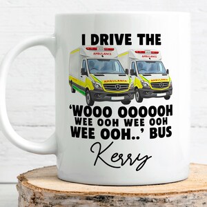 May include: A white ceramic mug featuring two yellow and white ambulance illustrations. The text reads: I DRIVE THE 'WOOO OOOOOOH WEE OOH WEE OOH WEE OOH..' BUS and the name Kerry in a black script font below the ambulance graphics.