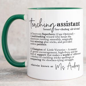 May include: A white ceramic mug with a dark green handle and interior. The mug features black printed text defining a teaching assistant as a classroom superhero and champion of little victories, ending with the personalised name Ms Audrey.