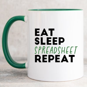 May include: A white ceramic mug with a dark green handle and interior. The front features the black text EAT, SLEEP, and REPEAT, with the word SPREADSHEET printed in green between them. The design is centred on the smooth, cylindrical surface.