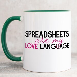 May include: A white ceramic mug with a dark green handle and interior rim. The front features the black text SPREADSHEETS and LANGUAGE, with the words are my LOVE in pink script lettering centred between the black lines of text.