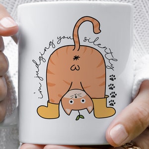 May include: White ceramic mug featuring a cartoon illustration of an orange cat wearing yellow boots and a green plant sprouting from its head. The cat is bent over with its tail in the air and the text "i'm judging you silently" is written on the mug.