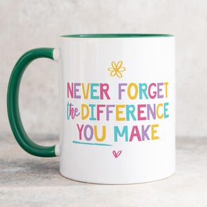 Könnte beinhalten: Ein weißer Keramikbecher mit dunkelgrünem Henkel und Innenrand. Auf der Vorderseite befindet sich ein gelbes Blumensymbol über dem mehrfarbigen Text NEVER FORGET THE DIFFERENCE YOU MAKE, mit einem kleinen rosa Herzsymbol unter dem Text.