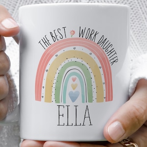 Può includere: Una tazza in ceramica bianca con una grafica arcobaleno dai colori pastello, decorata con piccoli cuori e un fiore. Il testo THE BEST WORK DAUGHTER è scritto sopra l'arcobaleno e ELLA sotto in un semplice carattere nero senza grazie.