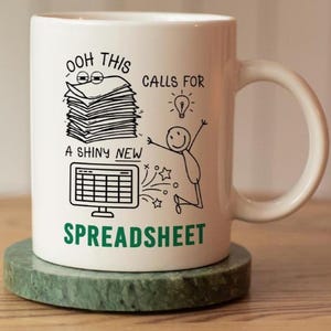 May include: A white ceramic mug featuring a black line drawing of a stack of papers, a computer monitor displaying a spreadsheet, a lightbulb, and a stick figure jumping. The text reads OOH THIS CALLS FOR A SHINY NEW SPREADSHEET in black and green.