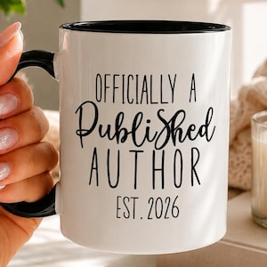 Könnte beinhalten: Ein weißer Keramikbecher mit schwarzem Innenraum und schwarzem Henkel. Auf der Vorderseite befindet sich der schwarze Schriftzug "OFFICIALLY A Published AUTHOR EST. 2026" in einer Kombination aus serifenloser Schrift und Schreibschrift.