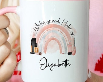 Può includere: Una tazza in ceramica bianca con manico e interno rosa. La parte anteriore presenta un grafico di un arcobaleno pastello con pennelli e flaconi per il trucco, il testo "Wake up and Make up" e il nome "Elizabeth" in corsivo nero.