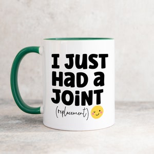 May include: A white ceramic mug with a dark green handle and interior rim. The front features bold black text that reads I JUST HAD A JOINT (replacement) followed by a small yellow winking smiley face emoji with pink cheeks.