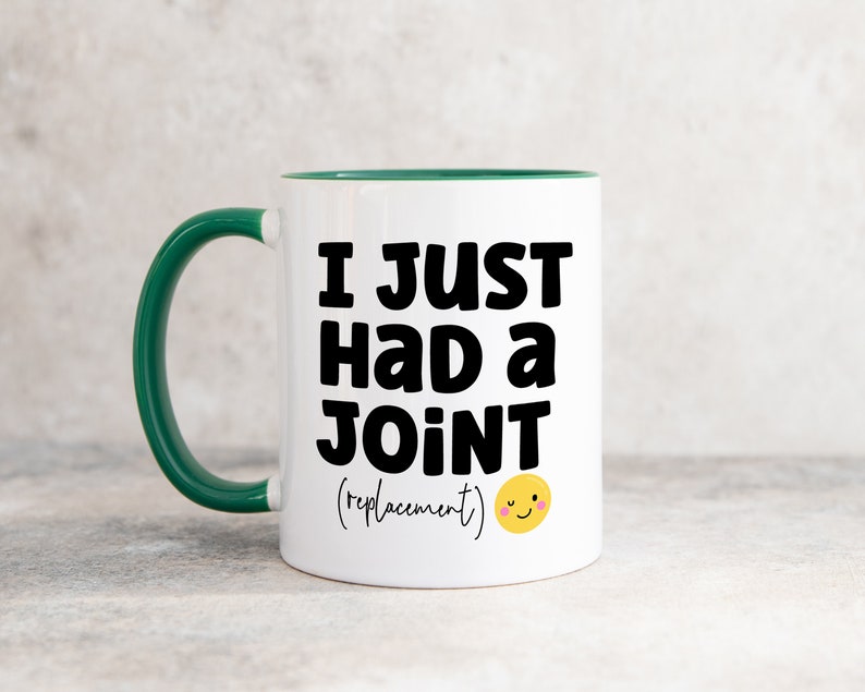 May include: A white ceramic mug with a dark green handle and interior rim. The front features bold black text that reads I JUST HAD A JOINT (replacement) followed by a small yellow winking smiley face emoji with pink cheeks.