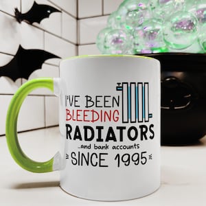 Op de afbeelding: Een witte keramische koffiemok met een groene handgreep. De mok heeft zwarte tekst die "I've Been Bleeding Radiators...and bank accounts since 1995" luidt.