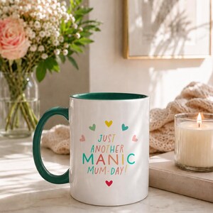 May include: A white ceramic mug with a dark green handle and interior. The front features the text "JUST ANOTHER MANIC MUM-DAY!" in colorful letters, surrounded by small, multi-colored heart icons arranged in a circular pattern.