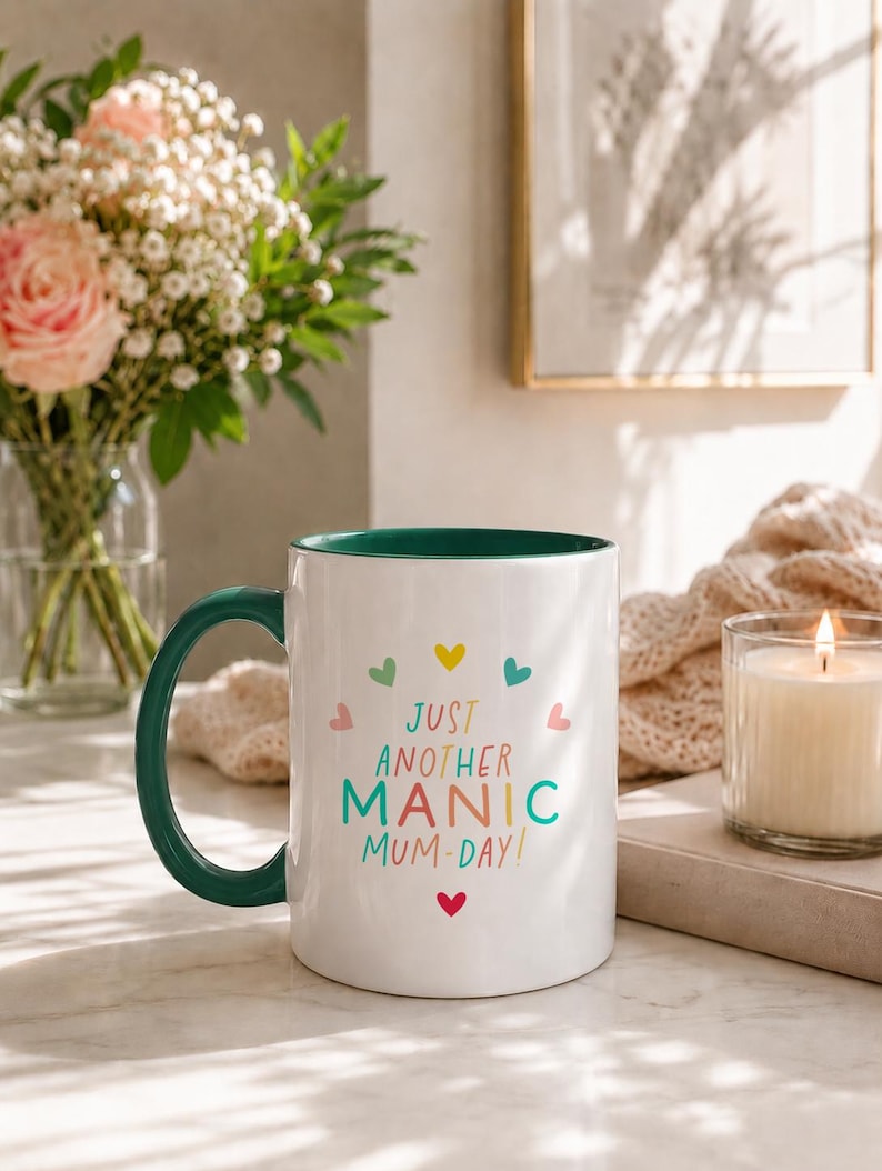 May include: A white ceramic mug with a dark green handle and interior. The front features the text "JUST ANOTHER MANIC MUM-DAY!" in colorful letters, surrounded by small, multi-colored heart icons arranged in a circular pattern.