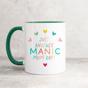 May include: A white ceramic mug with a dark green handle and interior. The front features the colorful text 'JUST ANOTHER MANIC MUM-DAY!' surrounded by small hearts in yellow, teal, pink, green, and red, arranged in a circular pattern.
