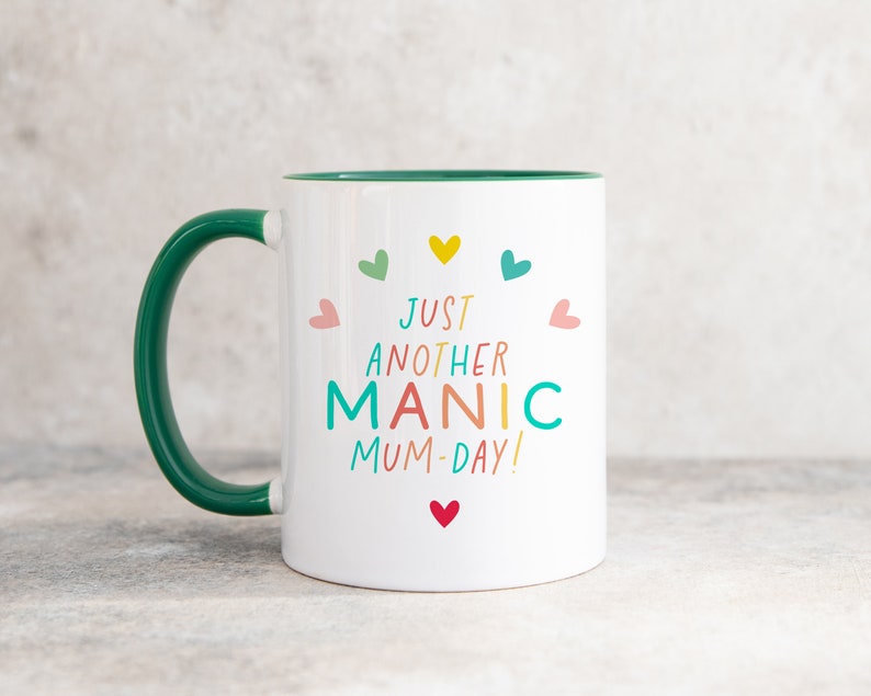 May include: A white ceramic mug with a dark green handle and interior. The front features the colorful text 'JUST ANOTHER MANIC MUM-DAY!' surrounded by small hearts in yellow, teal, pink, green, and red, arranged in a circular pattern.