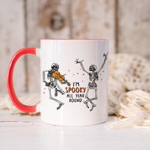 May include: White ceramic mug with a red rim and handle. The mug features a black and white illustration of two skeletons, one playing a violin. The text "I'm Spooky All Year Round" is printed on the mug.
