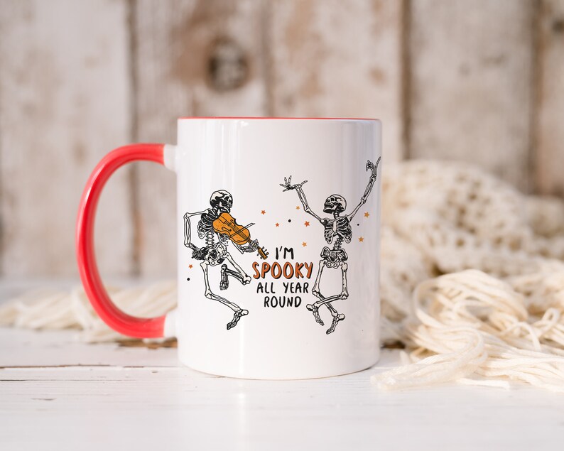 May include: White ceramic mug with a red rim and handle. The mug features a black and white illustration of two skeletons, one playing a violin. The text "I'm Spooky All Year Round" is printed on the mug.
