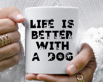 May include: A white ceramic mug featuring the black text "LIFE IS BETTER WITH A DOG" printed on the front. The letters are stylized with various silhouettes of dogs, paw prints, and bone shapes integrated into the typography design.
