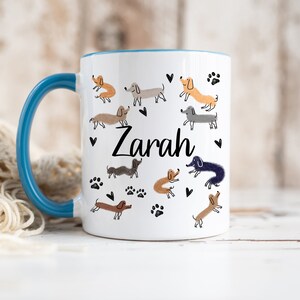 Pode incluir: Uma caneca de cer&acirc;mica branca com uma asa e rebordo interior azuis. O exterior apresenta um padr&atilde;o repetido de ilustra&ccedil;&otilde;es de c&atilde;es dachshund de v&aacute;rias cores, pegadas pretas e pequenos cora&ccedil;&otilde;es pretos. O nome Zarah est&aacute; impresso em preto.