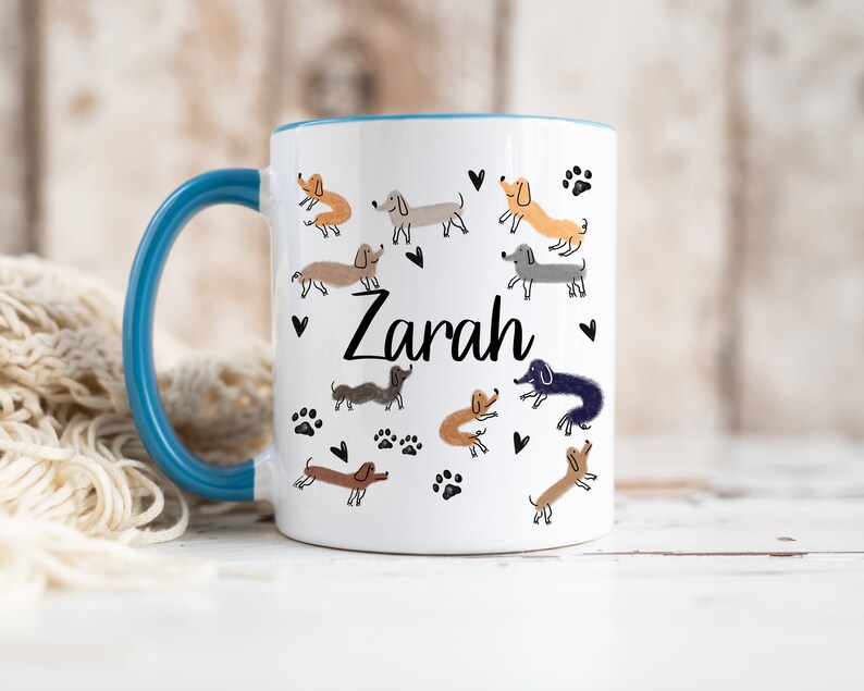 Pode incluir: Uma caneca de cer&acirc;mica branca com uma asa e rebordo interior azuis. O exterior apresenta um padr&atilde;o repetido de ilustra&ccedil;&otilde;es de c&atilde;es dachshund de v&aacute;rias cores, pegadas pretas e pequenos cora&ccedil;&otilde;es pretos. O nome Zarah est&aacute; impresso em preto.