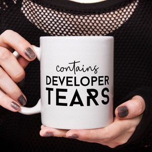 Peut inclure: Une tasse en céramique blanche avec du texte noir qui dit "contient des larmes de développeur".