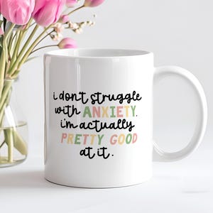 Peut inclure: Une tasse à café en céramique blanche avec un texte noir et coloré qui dit "Je ne lutte pas contre l'anxiété. En fait, je suis assez bon pour ça."