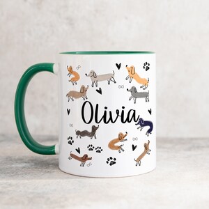 Pode incluir: Uma caneca de cer&acirc;mica branca com uma asa e rebordo interior verde-escuro. O exterior apresenta um padr&atilde;o repetitivo de c&atilde;es dachshund de v&aacute;rias cores, pegadas pretas, pequenos cora&ccedil;&otilde;es e ossos, com o nome Olivia em letra preta.